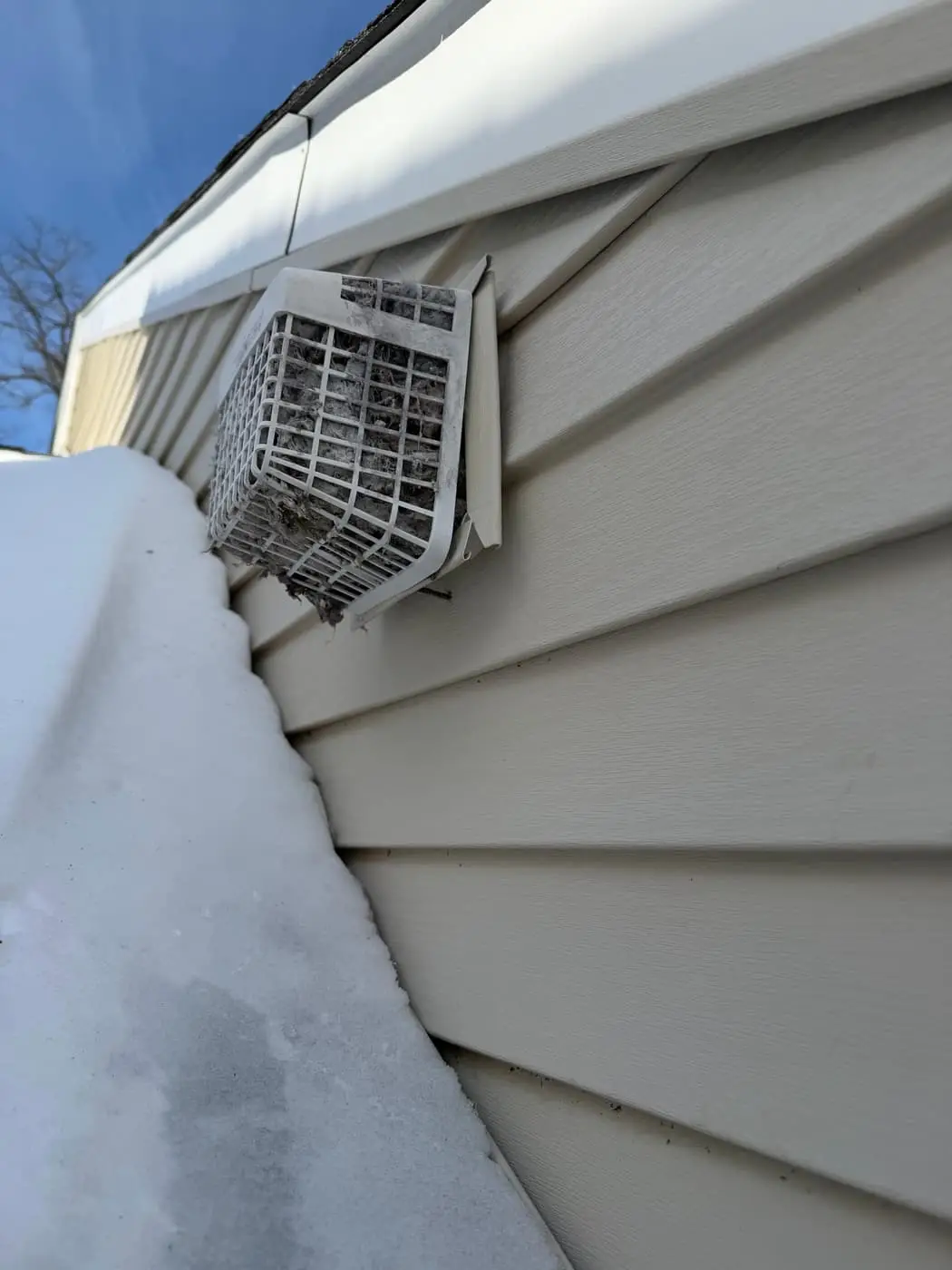 A dryer vent hood on vinyl siding with its louvers packed with lint, above a roof covered in snow.