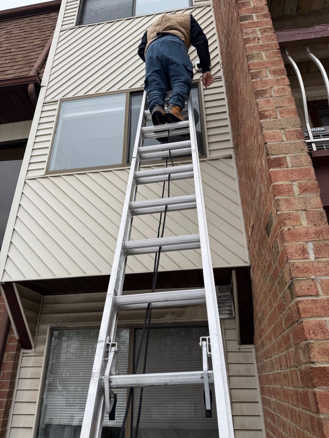 A technician climbing an aluminum extension ladder against a building with beige siding, beside a red brick wall.